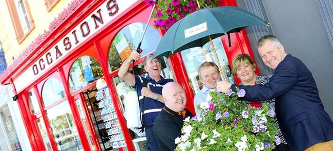 Group of people sheltering under an umbrella outside a red-fronted Waterford shopfront at a Waterford Business Group event