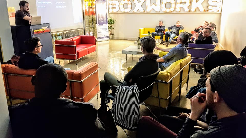 Audience seated on colourful chairs listening to a presentation at BOXWORKS beneath the illuminated BOXWORKS sign